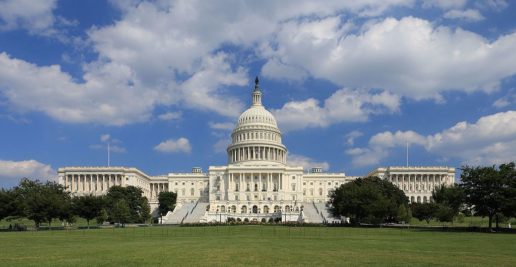 A photograph of the front of the U.S. Capitol Building. The lawn is green, and the sky is blue with big, puffy clouds.
