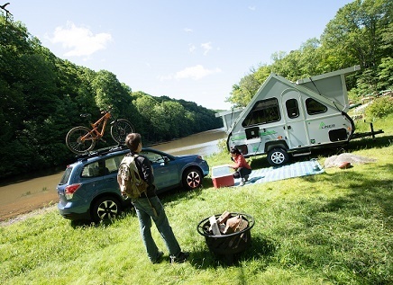 A photograph of a man standing next to a fire pit and a woman on a picnic blanket. Their SUV and pop-up travel trailer are parked behind them on a grassy river bank.Aliner camper lifestyle photo