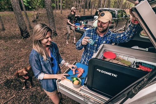 A woman cuts food on the back of a tailgate next to a Dometic CFX Cooler while a man leans against the vehicle and watches her. There is a brown dog behind the woman. Another man is sits on a different truck's tailgate in the background. The forest is all around them.