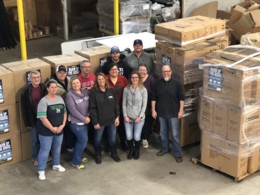 A phtograph of about a dozen people standing in front of pallets loaded high with boxes in a warehouse setting. Brent Stevens, general manager of Forest River’s XLR division (right) and members of his service group begin putting pallets of donated items together.