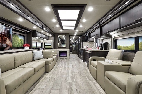 A photograph of the interior of a 2020 Thor Motor Coach Venetian. White leather couches and seats are on either side of the living room, and there is a fireplace against the wall on the far side of the kitchen.