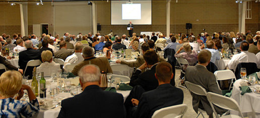 A photograph from the audience's perspective of a man standing at a podium addressing a conference hall full of people. This is the Recreational Vehicle and Motorhome Hall of Fame Induction Dinner