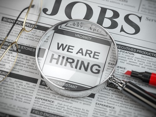 A picture of a newspaper with the word "Jobs" at the top of the page. A pair of reading glasses, a magnifying glass and a red marker all sit on top of the newspaper. The words "We Are Hiring" are visible under the magnifying glass.