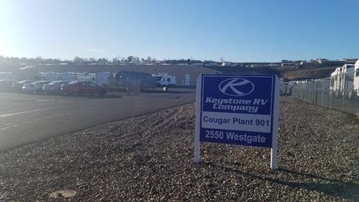 Photo of the exterior of the Keystone RV Plant in Oregon. A sign that reads "Keystone RV Company, Cougar Plant 901, 2550 Westgate" stands in a rock bed beside a parking lot full of cars and RVs. There is a warehouse at the far end of the parking lot.