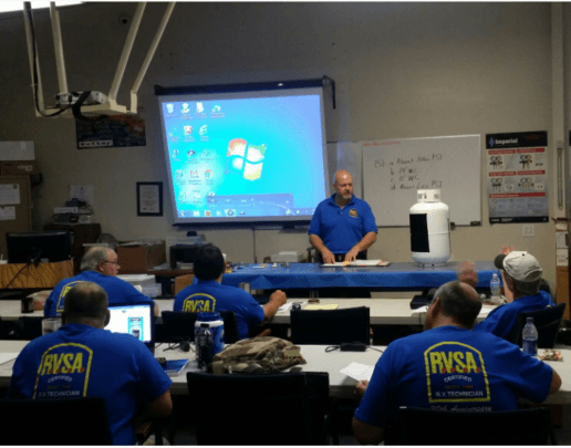 A picture of a man standing in front of a classroom of 5 students. The students are seated facing away from the camera and are all wearing blue RVSA t-shirts. The instructor is standing at a table with a propane tank on it. There is a projection screen and a white board on the wall behind him.