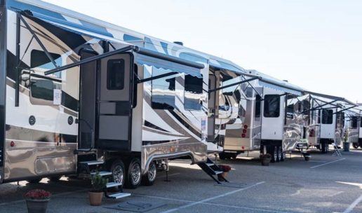 A row of clean, shiny travel trailers on a parking lot with their awnings out and doors open.