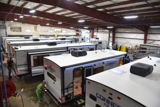 A photograph of about six KZ RV travel trailers lined up side-by-side in a manufacturing warehouse. There are a couple of employees working on the backs of different RVs.